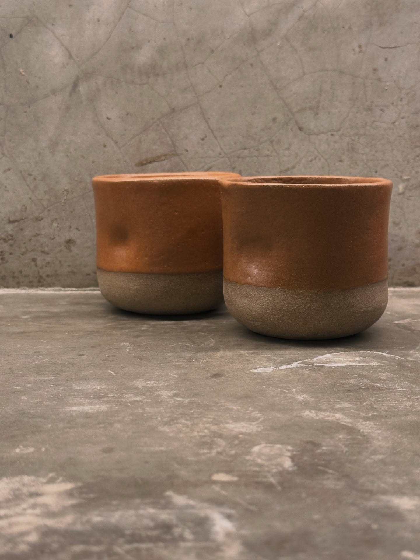 Dois copos de cerâmica terracota com base em barro cru, sobre superfície de cimento