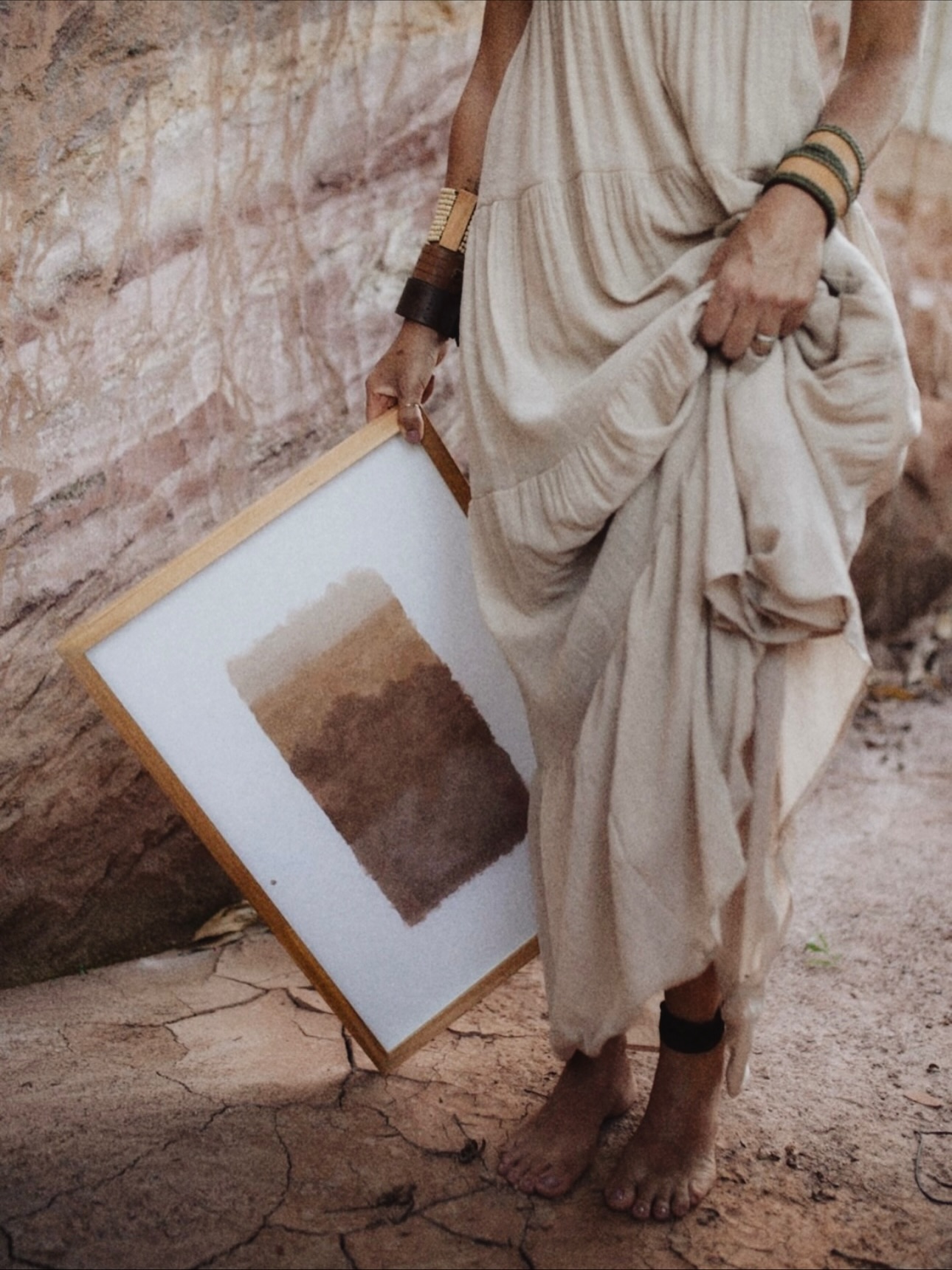 Dani descalça sobre a terra, segurando um quadro pintado com tintas de terra