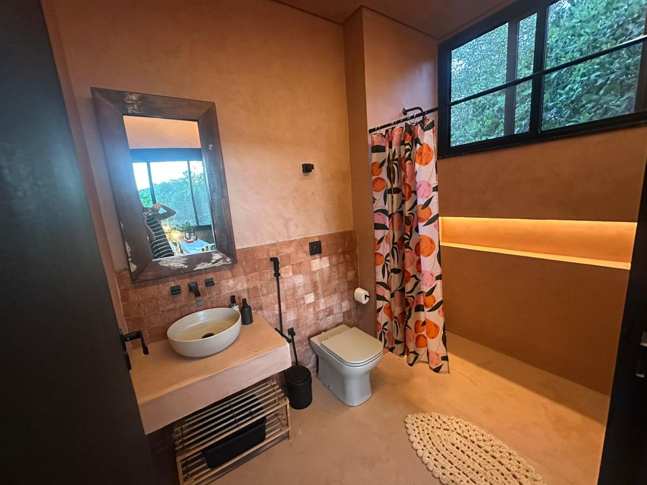 Banheiro em cimento queimado rosé, com tijolos de demolição, espelho de madeira, nicho iluminado e cortina estampada de cajus