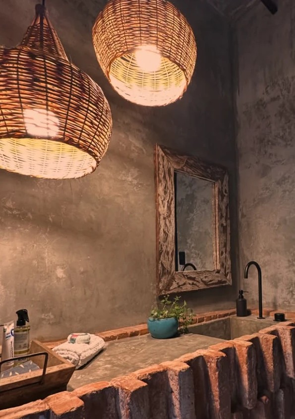 Lavabo com bancada de tijolos de demolição, paredes de cimento queimado, espelho de madeira e luminárias de palha